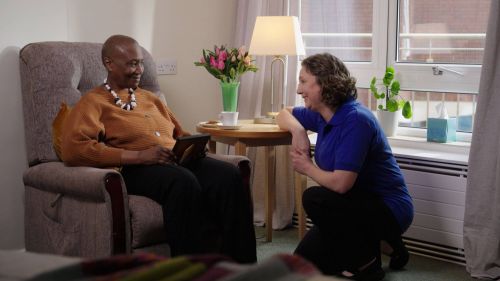 A caregiver kneels beside an older woman sitting in a comfortable armchair, smiling warmly at each other in a cozy bedroom in a care home with flowers and a lamp on a side table.
