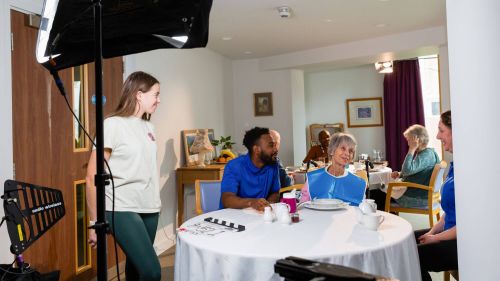 a behind the scenes shot of a camera assistant adjusting a light whilst smiling at actors sitting round a dining table