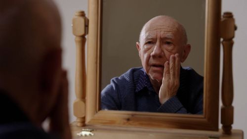 an older man looks at his reflection in the mirror, in his room in a care home, he is touching his face and looks confused